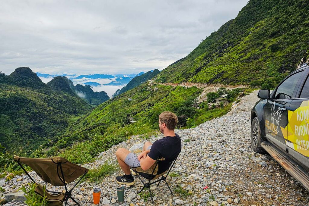 Ha Giang Loop By car with man sitting next to a car and Ha Giang's mountains as backdrop